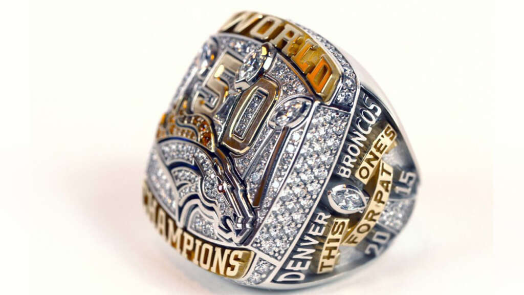 A championship ring with intricate details, featuring the number 50, a horse head emblem, and the words "World Champions," "Denver Broncos," "This One's For Pat," and "2015," embellished with numerous diamonds.