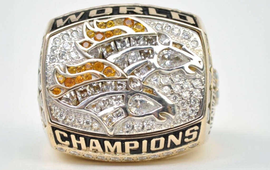 A championship ring adorned with the word "World Champions" and featuring two horse designs encrusted with diamonds and orange gems, symbolizing victory. The ring is set against a plain white background.