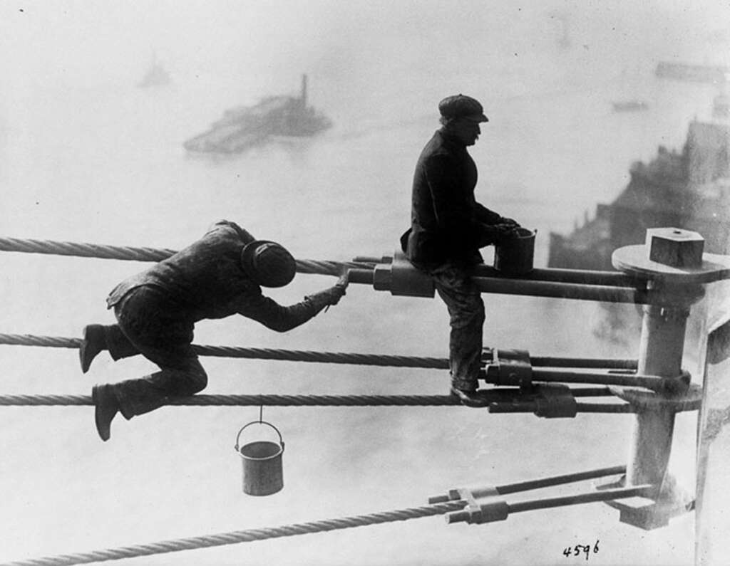 Two workers balancing on thick suspension cables of a bridge, high above a body of water. One man kneels, working with a tool, while the other stands holding a bucket. A foggy cityscape and boats are visible in the background.