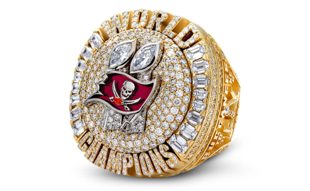 A gold Super Bowl championship ring encrusted with diamonds. The ring features the Tampa Bay Buccaneers logo, a pirate flag with a skull and crossed swords, set against a sea of small diamonds. The words "World Champions" encircle the design.