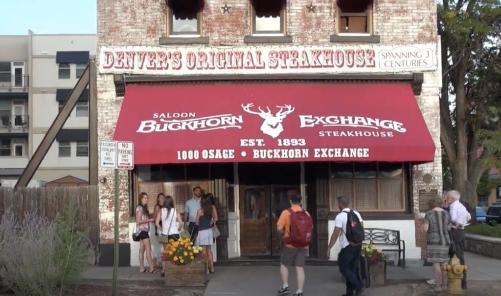 A group of people stands outside the historic Buckhorn Exchange Steakhouse in Denver. The building is rustic with a red awning displaying the name and "Est. 1893." A sign reads "Denver's Original Steakhouse" and "Spanning 3 Centuries.