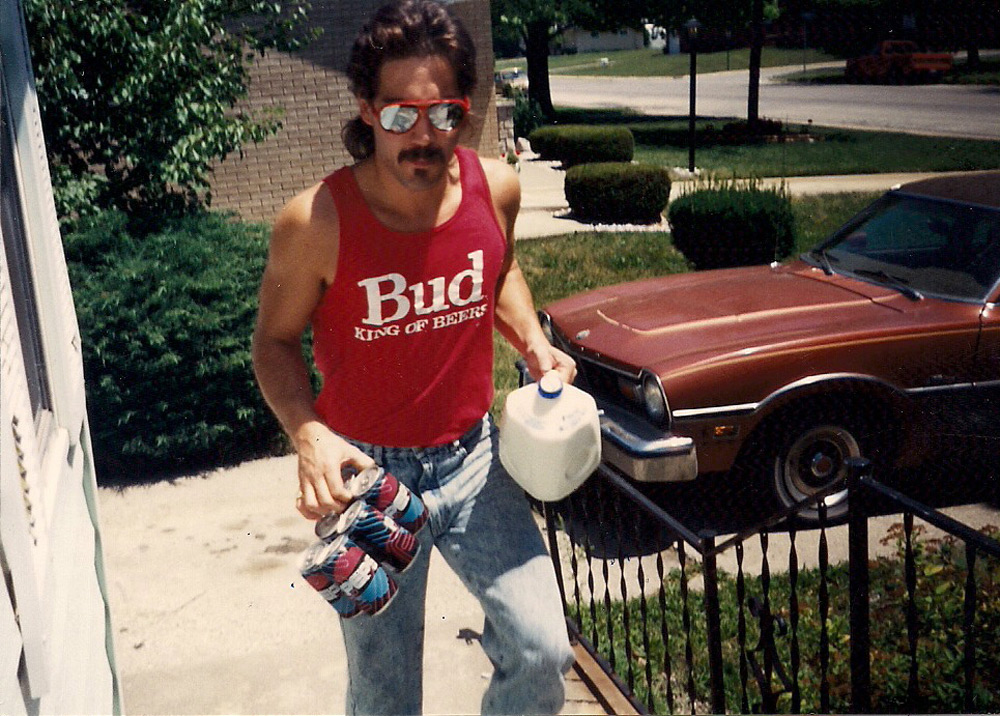 A man in sunglasses and a red "Bud" tank top carries a gallon of milk and a six-pack of beer bottles while walking up outdoor steps. A brown car and a lawn are in the background.