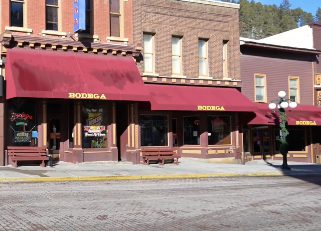 Street view of a brick building with red awnings labeled "Bodega." Large windows display signs. The sidewalk is bordered by a weathered bench and a decorative lamp post.