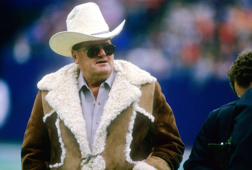 Man wearing a large white cowboy hat, sunglasses, and a brown shearling coat stands outdoors at a sporting event. The background is blurred, showing a crowd of people.