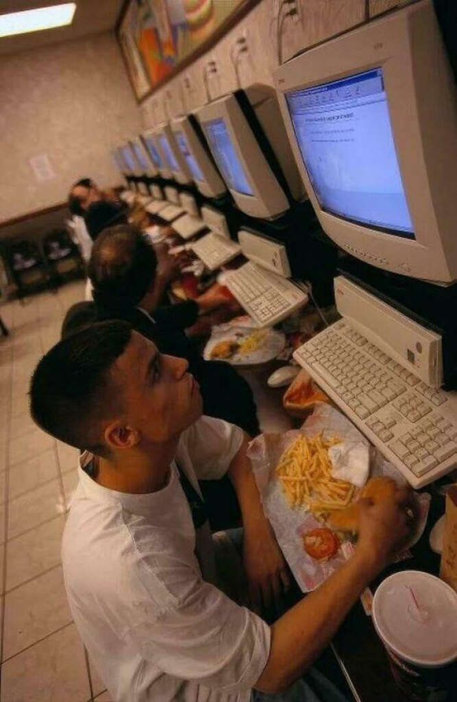 A row of people sitting in front of old desktop computers in a cafe. The foreground shows a person in a white shirt eating fries with a sandwich while using the computer. Other patrons are similarly focused on their screens.