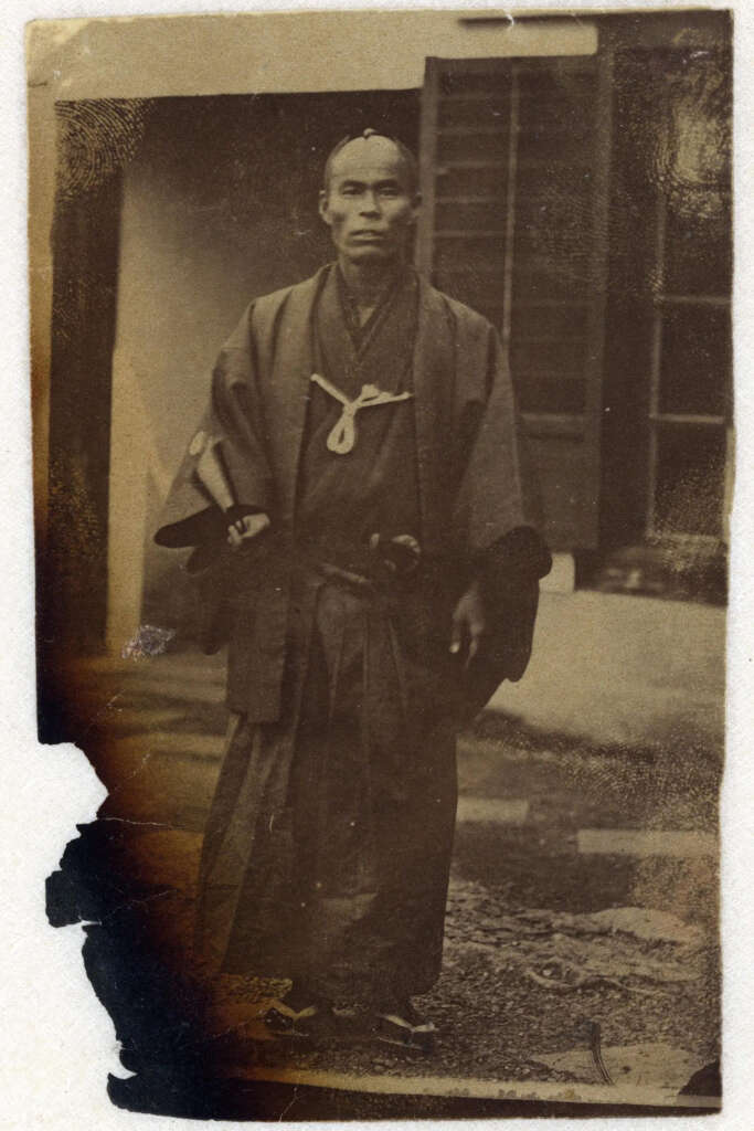 Sepia-toned vintage photo of a man in traditional Japanese attire. He wears a kimono and hakama, holding a sheathed sword. The background features a building with an open door. The photo shows signs of age and wear.