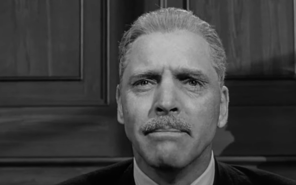 Black and white image of a man with a serious expression, short light-colored hair, and a mustache. He is wearing a suit and is pictured against a wooden background, likely in an indoor setting.