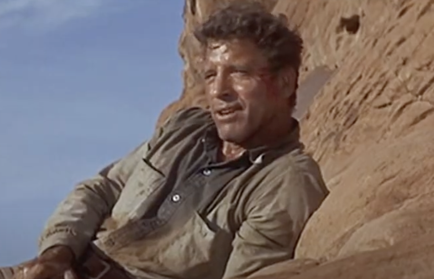 A man in a rugged, dusty shirt leans against a rocky surface. His expression is determined, and the background shows a clear blue sky.