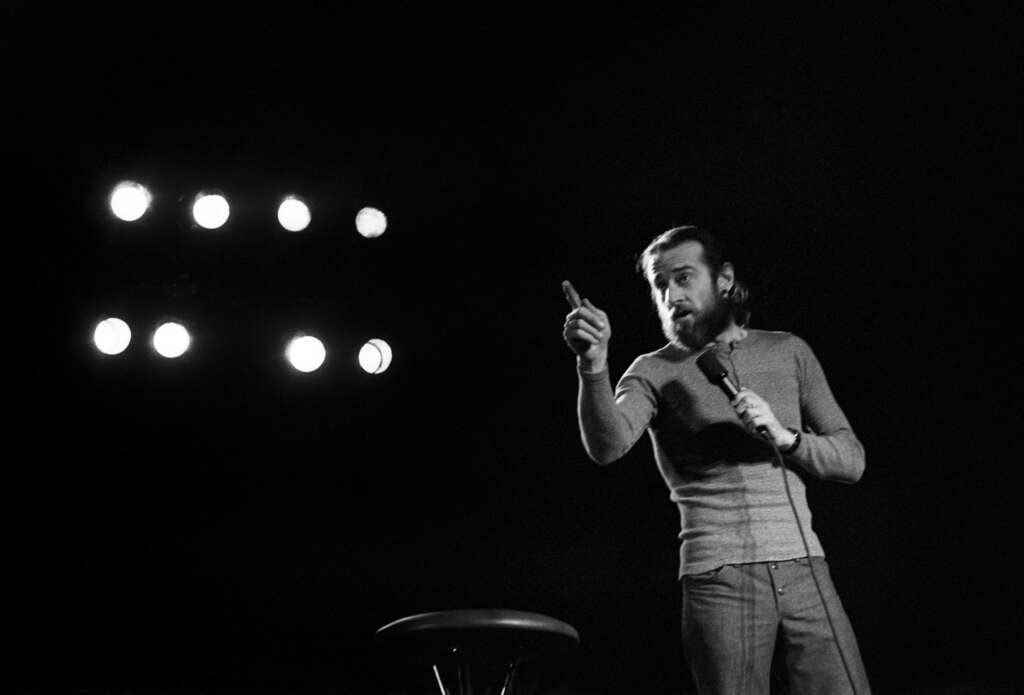 A bearded man stands on stage with a microphone, gesturing animatedly with his right hand. He wears a long-sleeve shirt and pants. Bright stage lights are visible in the background, and a stool is beside him.