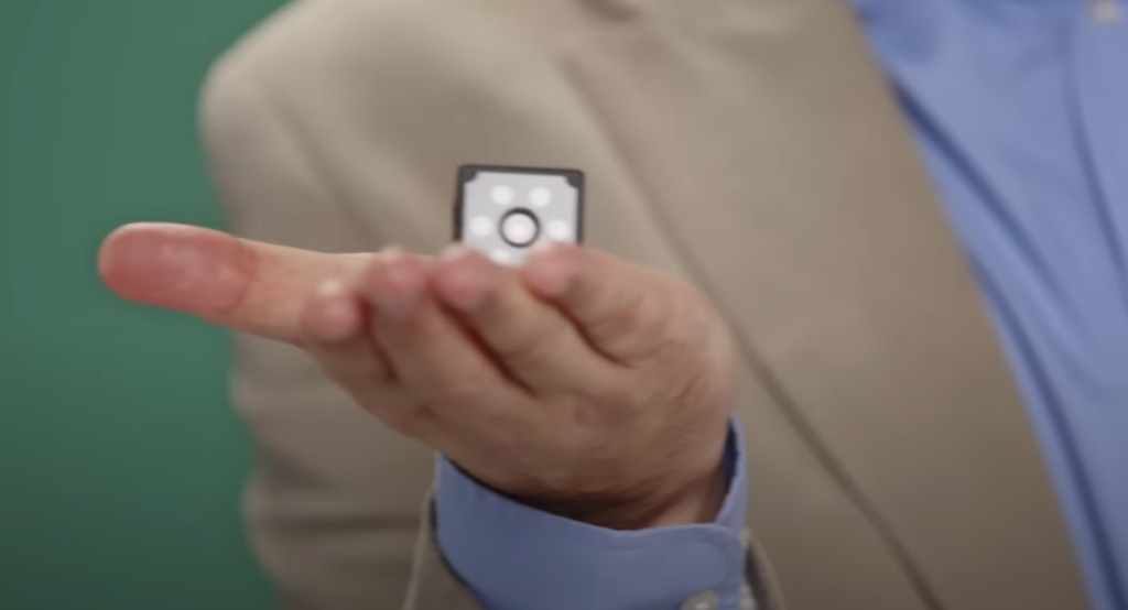 A person in a beige blazer and blue shirt holds a small black and silver object on their open palm. The object appears to have a black circle and small white shapes on its surface. The background is green.