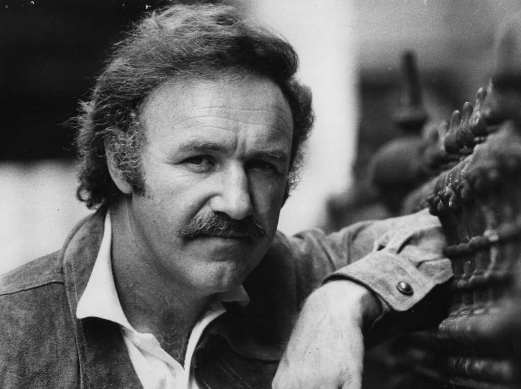 A black-and-white portrait of a middle-aged man with wavy hair and a mustache, wearing a collared shirt and a jacket. He rests his arm on a textured surface, looking directly at the camera with a serious expression.