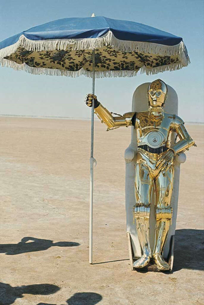 A golden humanoid robot sits under a blue and patterned beach umbrella in a desert landscape. The robot is reclined on a white chair, with a vast open expanse and clear sky in the background. Shadows are cast on the ground.