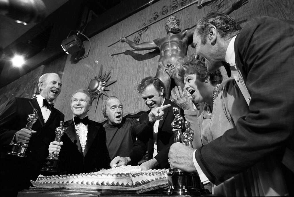 A group of six people is gathered around a large cake. Some are holding Oscar statues. In the background, there's a wooden wall with a sculpture of a man with outstretched arms. They appear to be celebrating, smiling and talking.