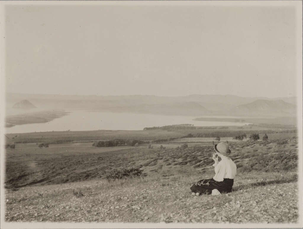 A person sits on a hillside overlooking a vast landscape with a large body of water in the distance. They are wearing a hat and facing the view. The scene is calm, with rolling hills under a clear sky.