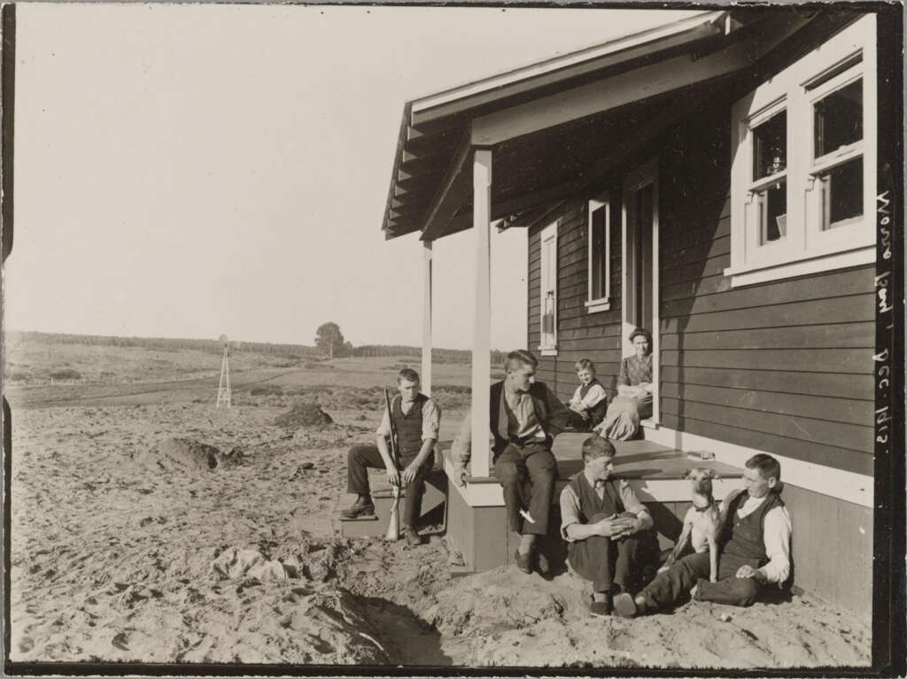 A black-and-white photo of six people sitting on the steps of a wooden house in a rural area. The landscape features open fields and a windmill. The group includes men, women, and children, all casually dressed, enjoying a sunny day.