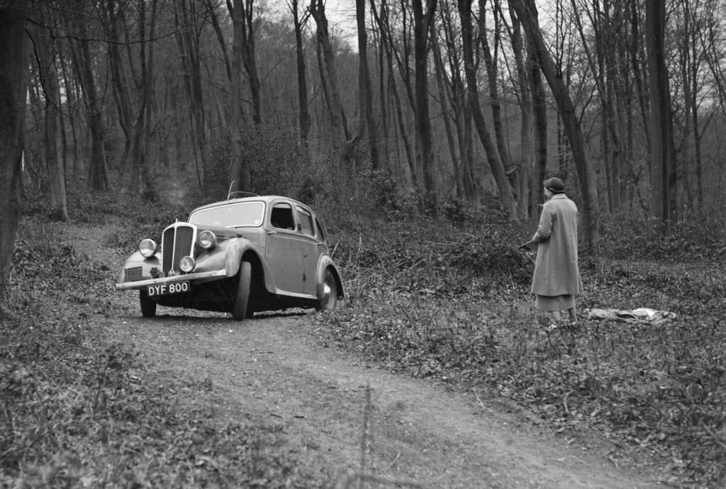 Black and white photo of a vintage car stuck on a muddy forest path. A person in a coat stands nearby holding a spade, possibly preparing to help. Leafless trees surround the scene, adding to the cold, wintry atmosphere.