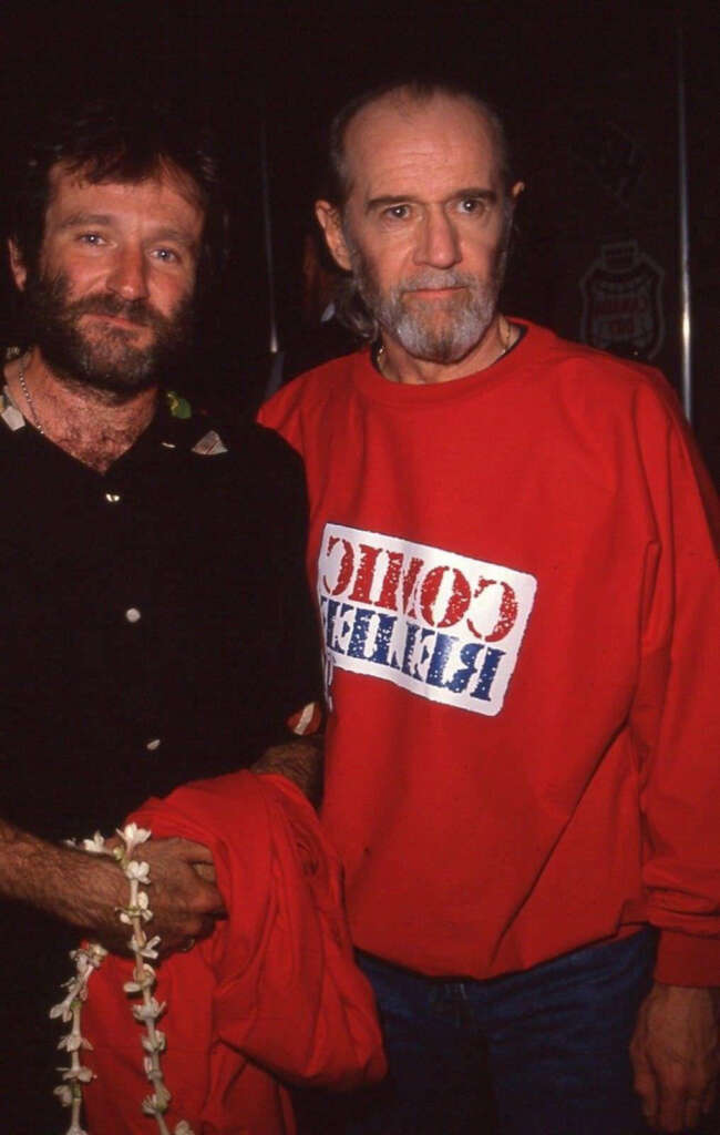 Two men are standing side by side. The man on the left has a beard and is wearing a black shirt, holding a red jacket and a white lei. The man on the right is wearing a red sweatshirt with a partially visible graphic design.