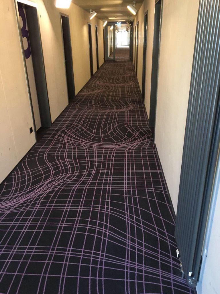 A long corridor with a patterned carpet featuring wavy, purple lines on a dark background. Doors line each side, and lights are mounted on the walls, leading to a bright area at the end of the hallway.