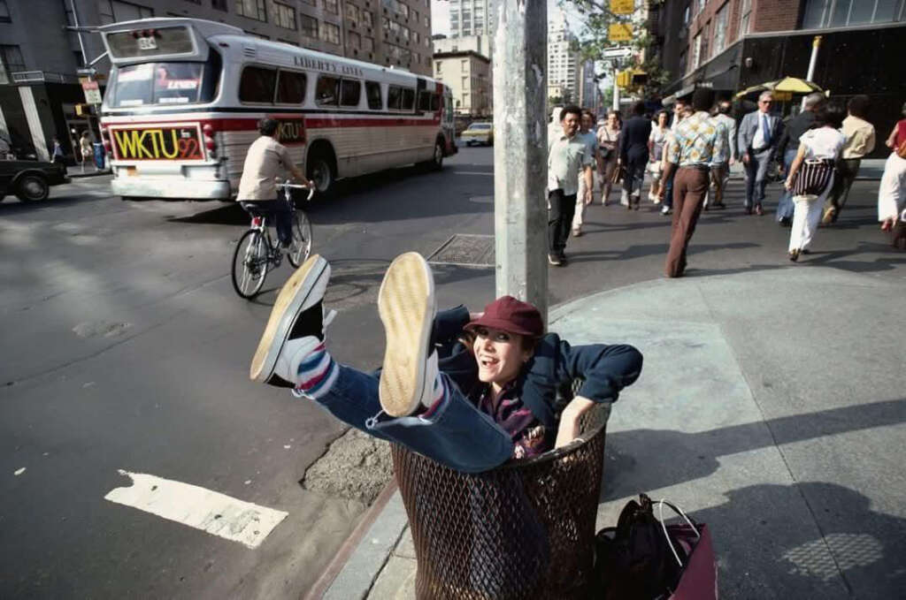 Person playfully sitting in a street trash bin with legs up, wearing a hat and sneakers. A cyclist passes by, and a bus drives on the street. Pedestrians walk on the sidewalk in the background, with tall buildings lining the street.