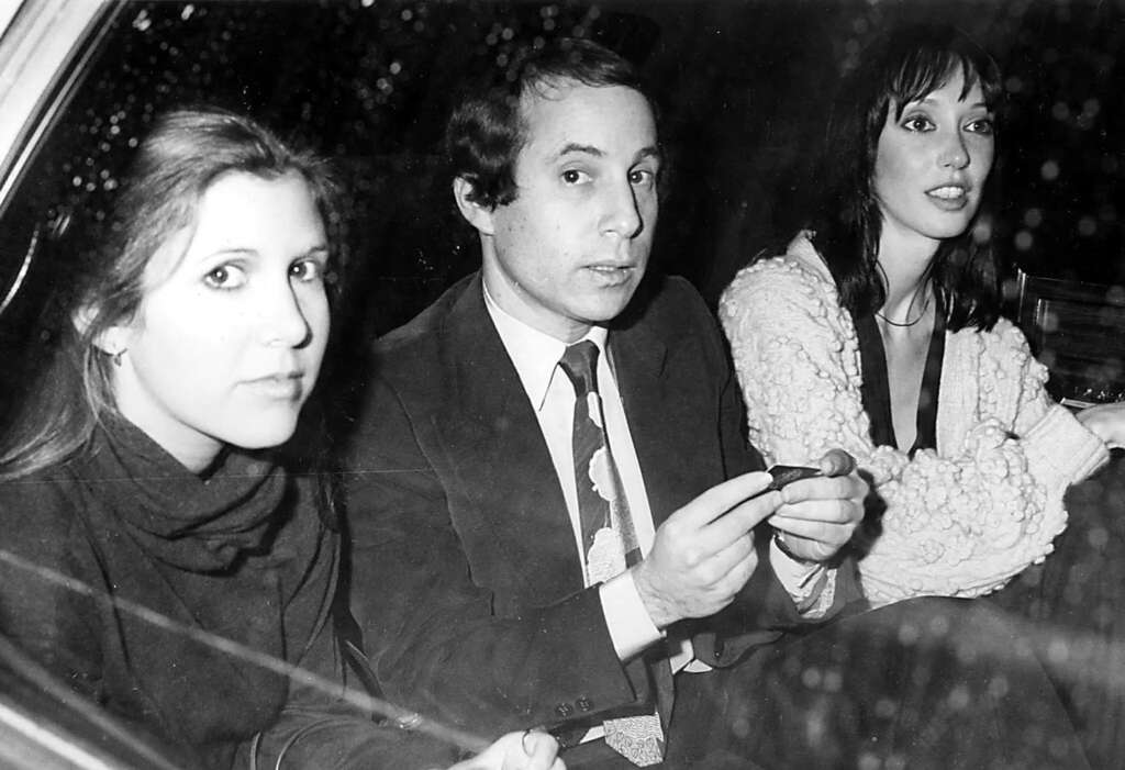 A black and white photo of three people sitting in the back of a car. A woman on the left wears a dark top, the man in the center holds a cigar, and a woman on the right wears a light-colored, textured jacket. The window is speckled with raindrops.