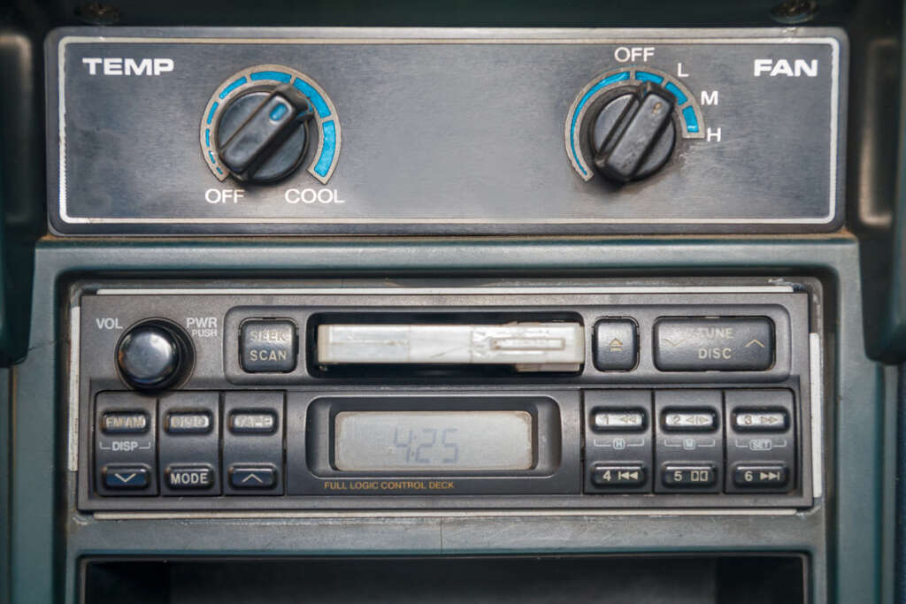 Close-up of a vintage car stereo system with an integrated cassette player and digital clock showing 4:25. Above it, there are two round dials labeled TEMP and FAN, used for controlling air conditioning settings.