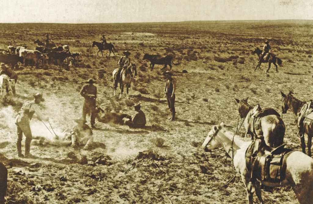 A sepia-toned image depicts cowboys on horseback in a barren landscape. Several men are gathered around a campfire, while others look on or ride horses. Sparse vegetation and open plains stretch into the distance under a clear sky.