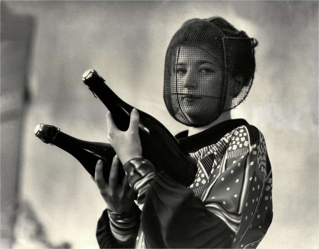 A woman with a mesh face covering holds two large bottles in her arms. She is wearing a patterned outfit and has her hair styled up. The background is blurred, putting focus on her and the bottles. The image is in black and white.