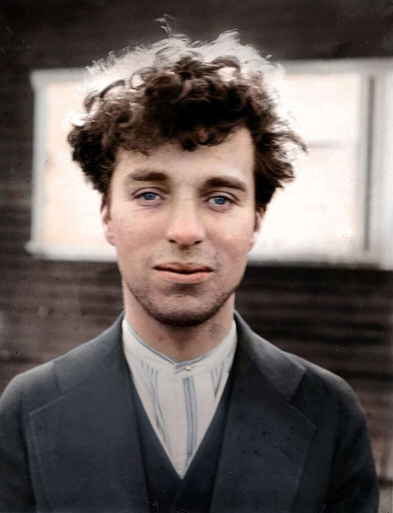 A man with curly hair and blue eyes wears a dark suit and striped shirt, looking directly at the camera. The background is blurred, suggesting an outdoor setting with a building behind him. The image has a vintage, colorized look.