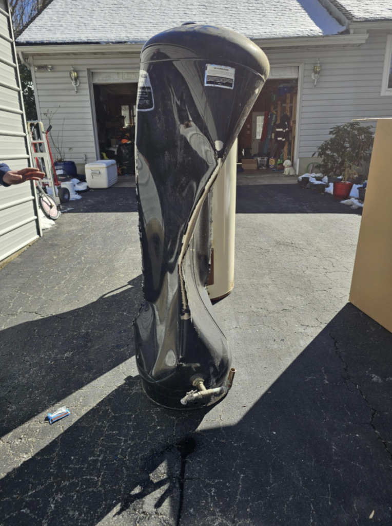 A twisted and damaged water heater stands upright on a driveway. The heater is black, with visible dents and creases. It is positioned in front of a garage, with various items scattered around the area.