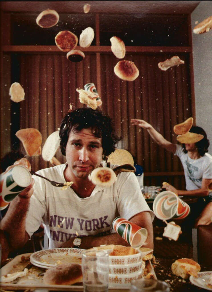 A person wearing a "New York" t-shirt is sitting at a table with a cluttered plate. Various bread items and cups are suspended mid-air around him. Another person in the background seems to be tossing something.