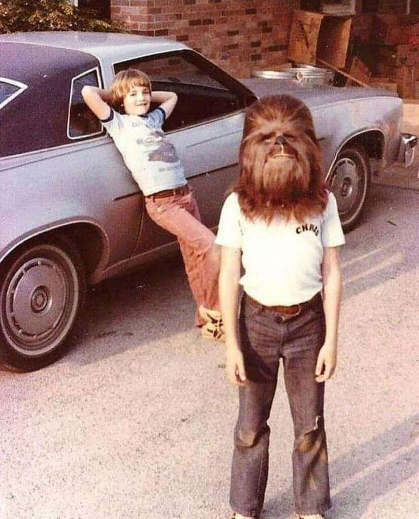 A child wearing a Chewbacca mask and a "CHRIS" shirt stands in front of a vintage car. Another child leans against the car, smiling, in a casual pose. The setting appears to be a residential driveway.