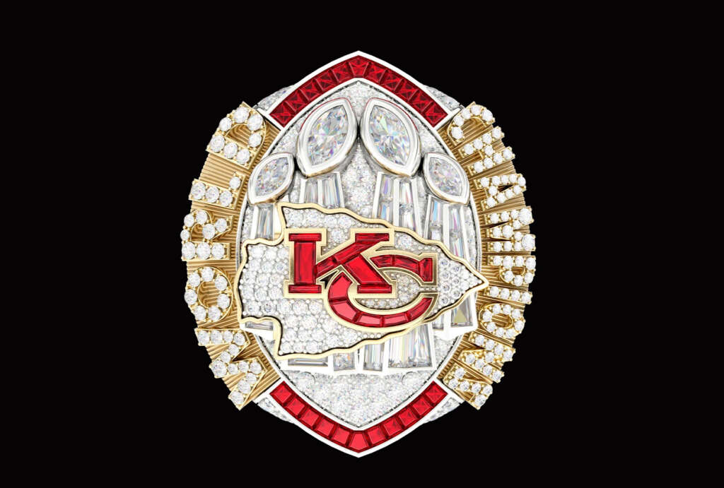 A championship ring with a prominent red and gold team logo in the center, surrounded by numerous diamonds and gemstones. The edges feature intricate designs with more jewels, set against a black background.