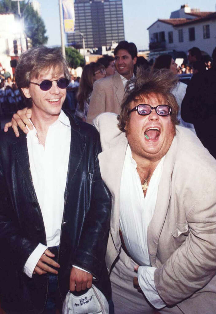 Two men are smiling and posing playfully at an outdoor event. The man on the left wears sunglasses and a leather jacket, while the man on the right wears glasses and a light-colored suit, making a humorous expression. People and buildings are in the background.