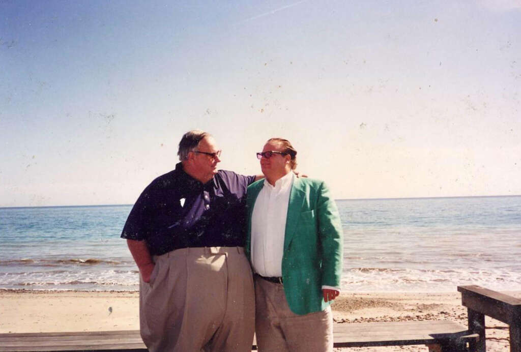 Two men stand together on a beach, looking at each other. One wears a dark shirt and beige pants, while the other wears a green jacket and white shirt. The ocean and clear sky are visible in the background.