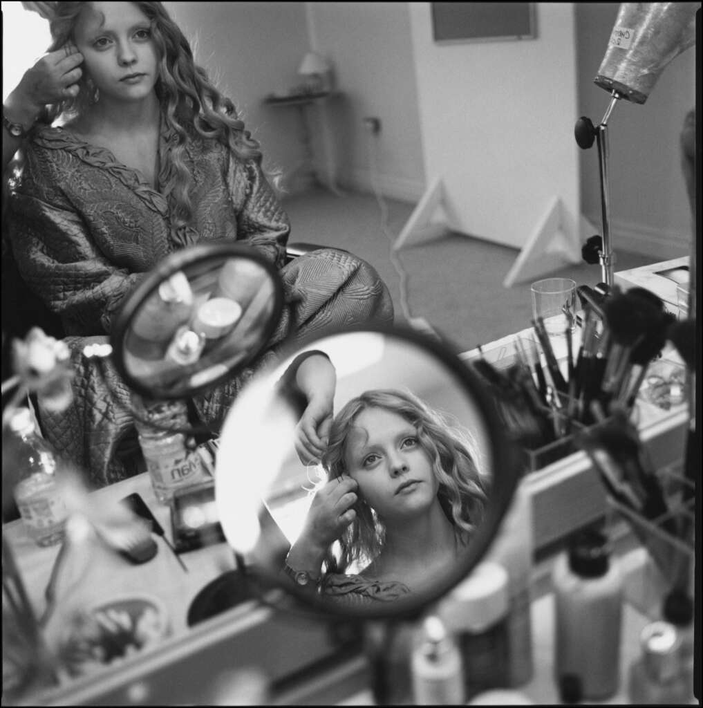 A person with curly hair sits at a reflective makeup table, surrounded by cosmetics and tools. They wear a patterned robe and are looking at their reflection in a small circular mirror, while someone adjusts their hair.