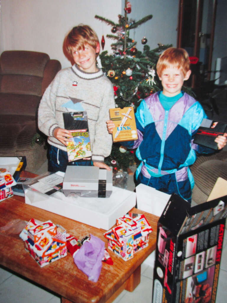 Two children smiling while holding video game boxes in a living room with a Christmas tree in the background. A table is in front of them with wrapped gifts and an open Nintendo box.