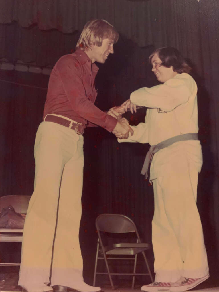 Two people are shaking hands on a stage. One, wearing a red shirt and white pants, stands smiling. The other, dressed in a martial arts uniform with a colored belt, holds a certificate. Two chairs are visible in the background.