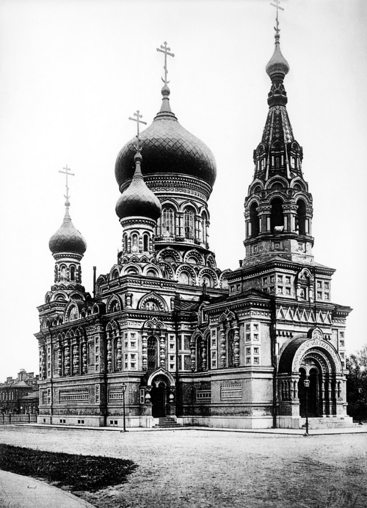 A black and white photograph of a Russian Orthodox church with large onion domes and intricate architectural details, including arched windows, ornate decorations, and crosses atop the domes. The sky is clear, and the ground is bare.