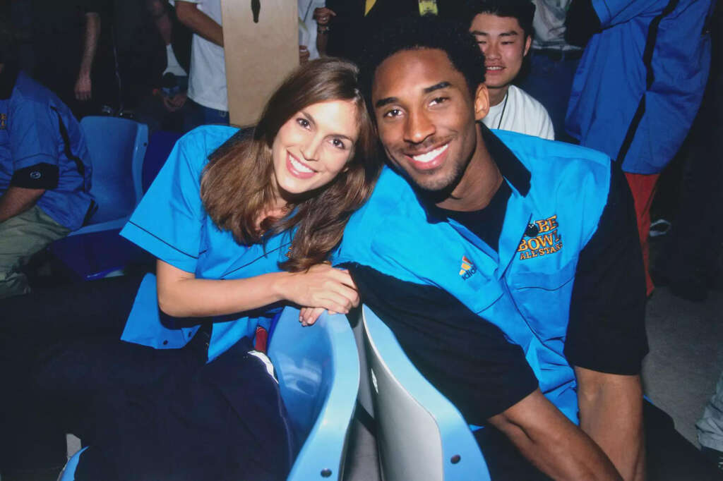 A man and a woman are sitting closely together on blue chairs, both wearing blue shirts with logos. They are smiling, and several other people are visible in the background. The setting appears to be a casual or event environment.