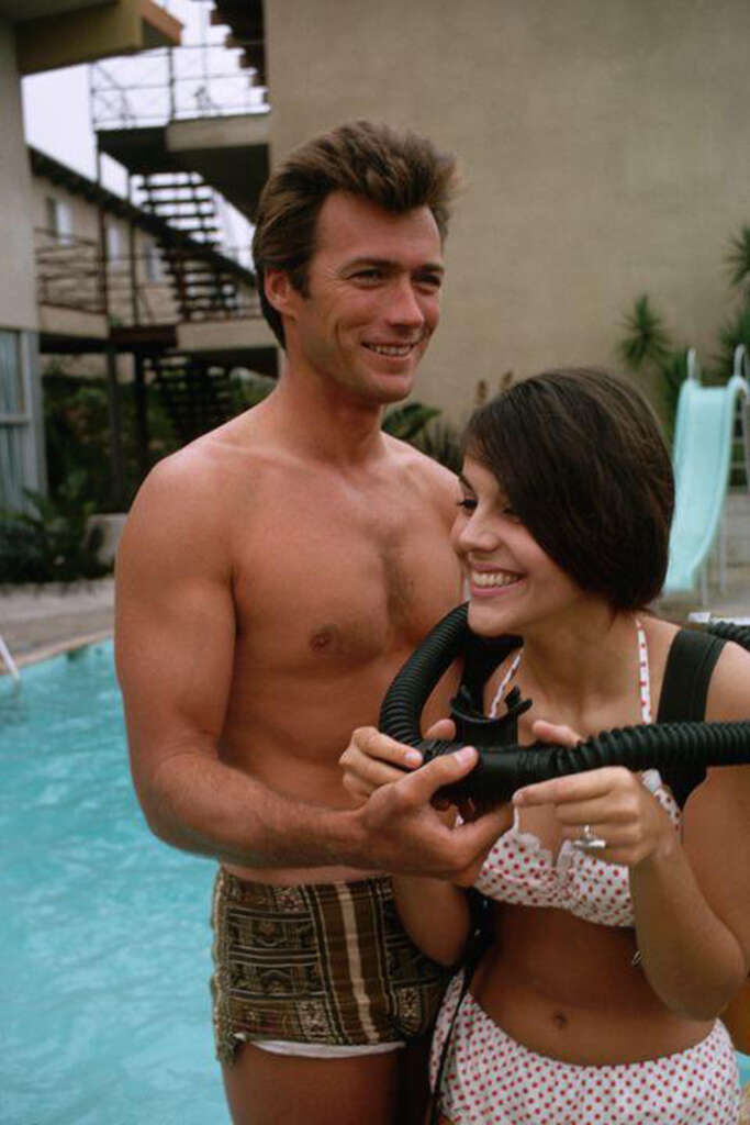 A shirtless man and a woman in a polka dot swimsuit stand by a pool. The man smiles, looking forward, while the woman holds a snorkel mask, laughing. A slide and building with outdoor stairs are visible in the background.