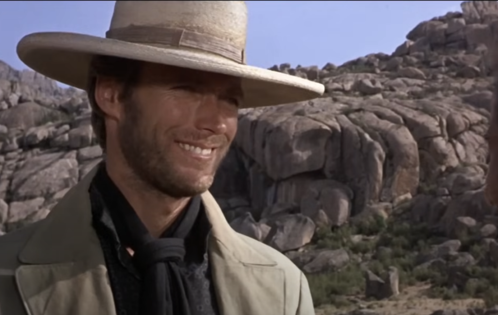 A person in a wide-brimmed hat and light-colored coat stands outdoors in a rocky landscape, smiling faintly. The background features large rock formations under a clear blue sky.