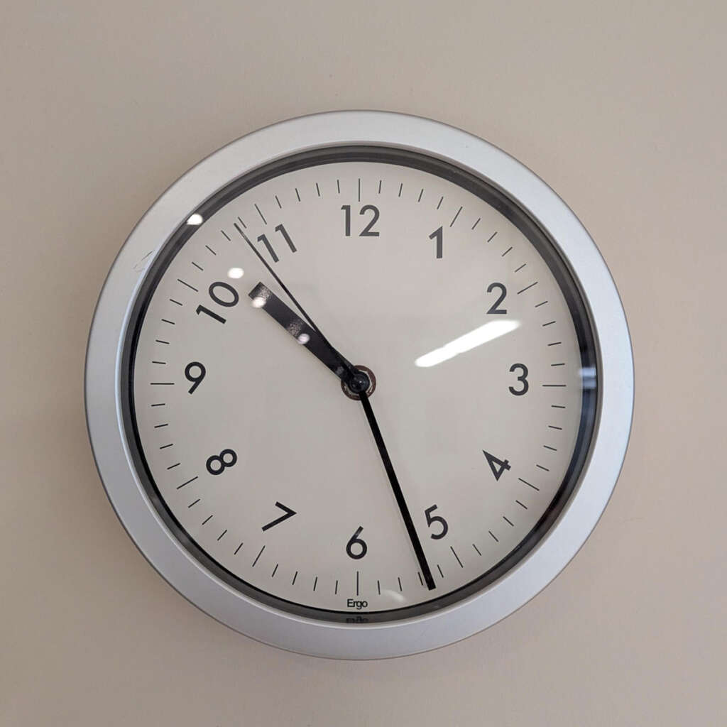 A round wall clock with a silver frame showing the time as 10:11. The clock face has black numbers and hands against a white background. A reflection of light is visible on the clock's glass surface.