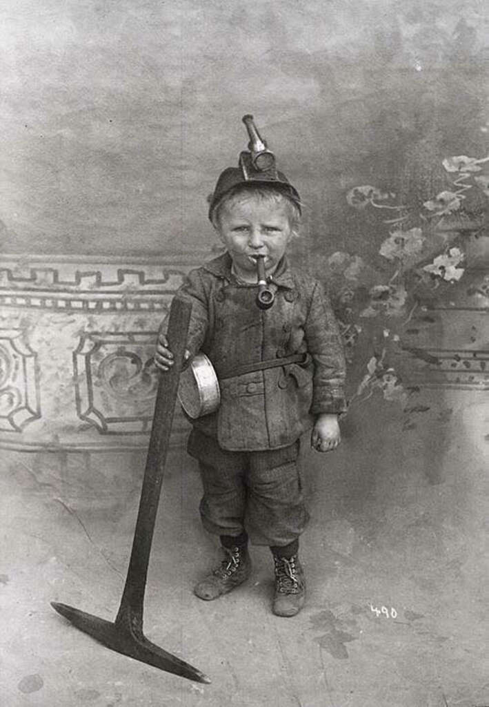 A young child dressed as a coal miner stands holding a mining tool. The child wears a cap with a lantern and a dusty outfit, with a pipe in their mouth. The backdrop features a decorative design. The photo is in black and white.