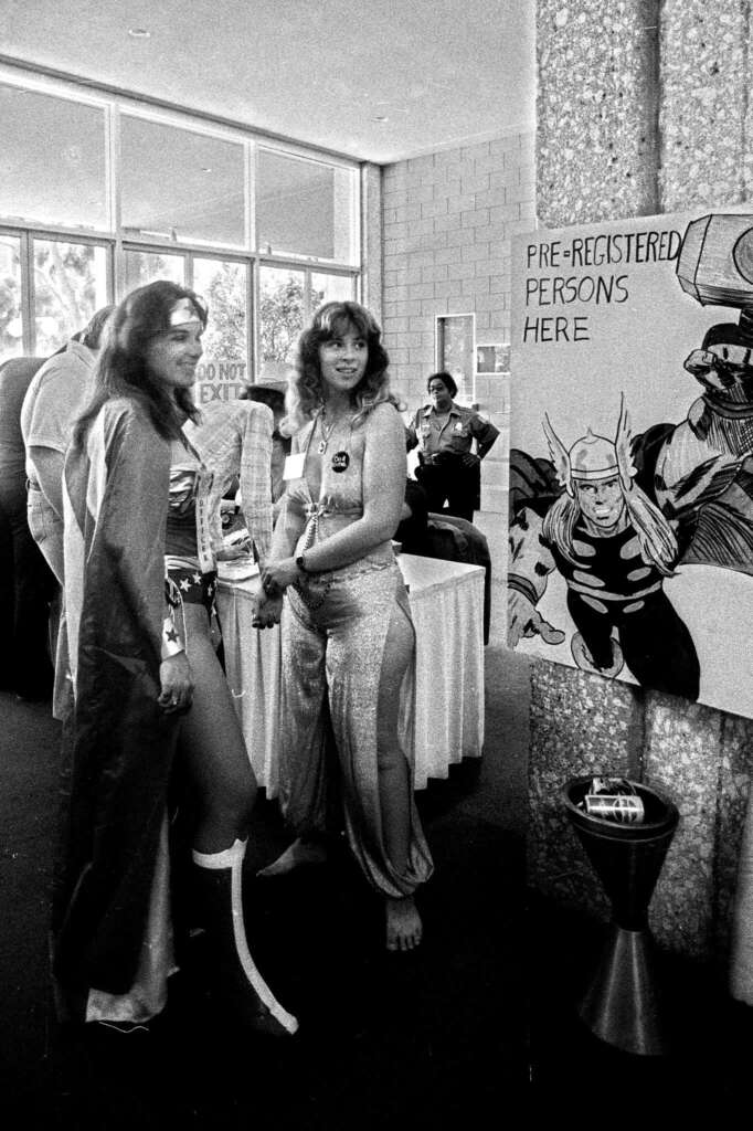 Two people in costume stand in a convention setting. One wears a cape and boots, the other a shiny outfit. A sign reads "Pre-registered persons here" with art of a superhero. Attendees mill about in the background.