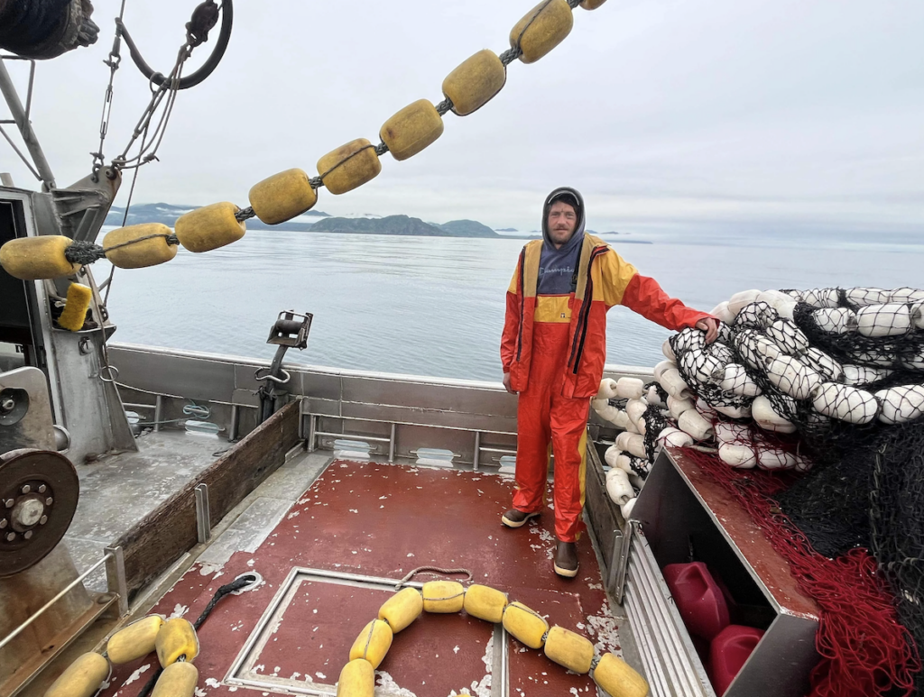 A person in orange and yellow rain gear stands on a fishing boat's deck. They are surrounded by ropes and fishing nets with floats. The ocean and distant mountains are visible in the background under a cloudy sky.