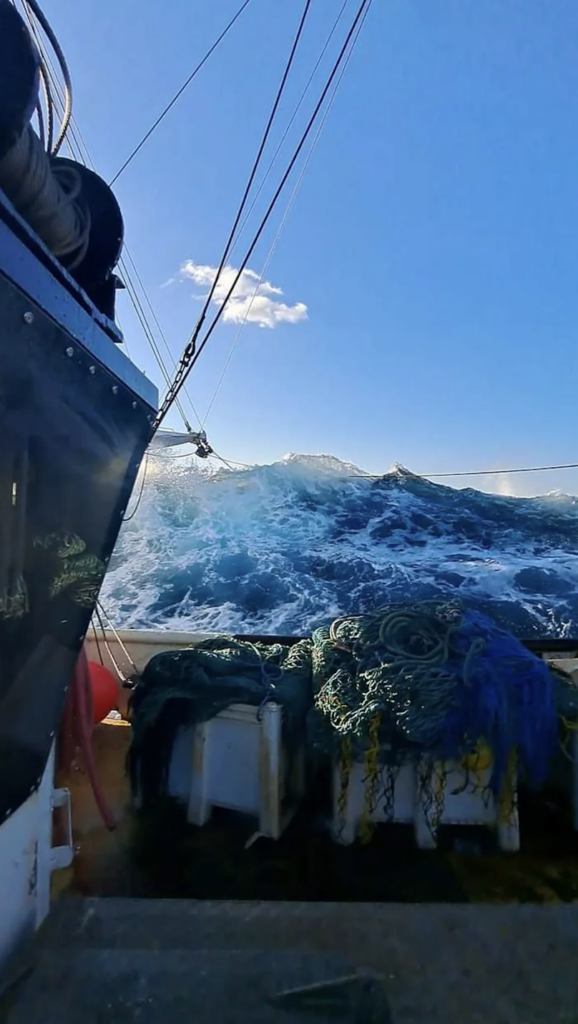 A fishing boat is on turbulent seas with a large wave towering in the background. The deck is cluttered with fishing nets and equipment. The sky is bright blue with a few clouds.