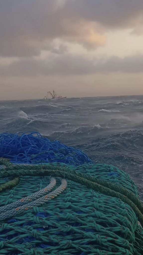 A turbulent sea with big waves dominates the foreground, displaying blue and green fishing nets. In the background, a fishing boat is visible under a cloudy, overcast sky.