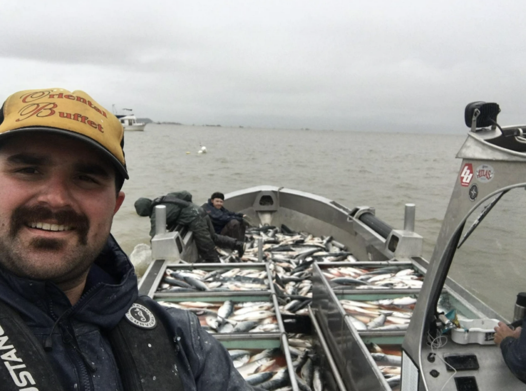 A person wearing a yellow cap and navy jacket is taking a selfie on a boat filled with numerous fish. Two other people can be seen handling the catch. Overcast sky and water are in the background.