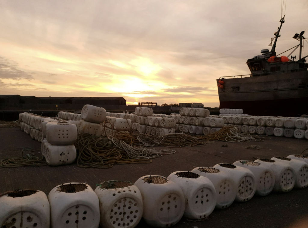 Sunset over a harbor with a docked boat. Numerous white, perforated cylindrical containers are stacked on the ground, alongside coiled ropes. The sky is filled with warm hues of orange and yellow.