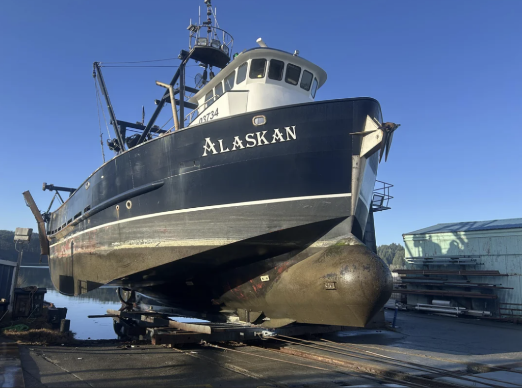 Fishing boat named "Alaskan" with a black hull and white upper section, elevated on a slipway in a shipyard. The environment is sunny, and the ship is clearly out of the water, showing the large underbelly and propellers.